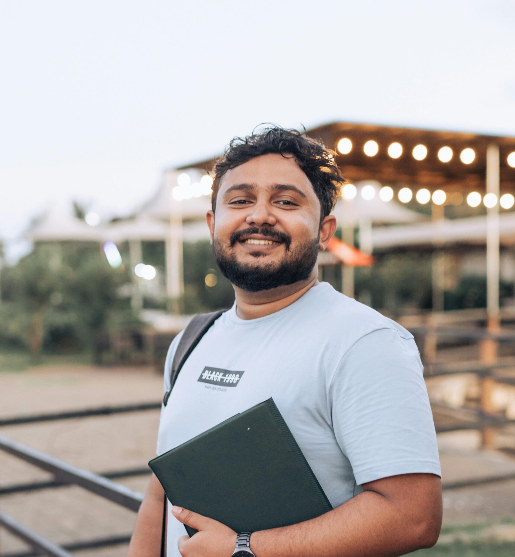 Cheerful man with a beard holding a notebook outdoors, dressed in casual attire.